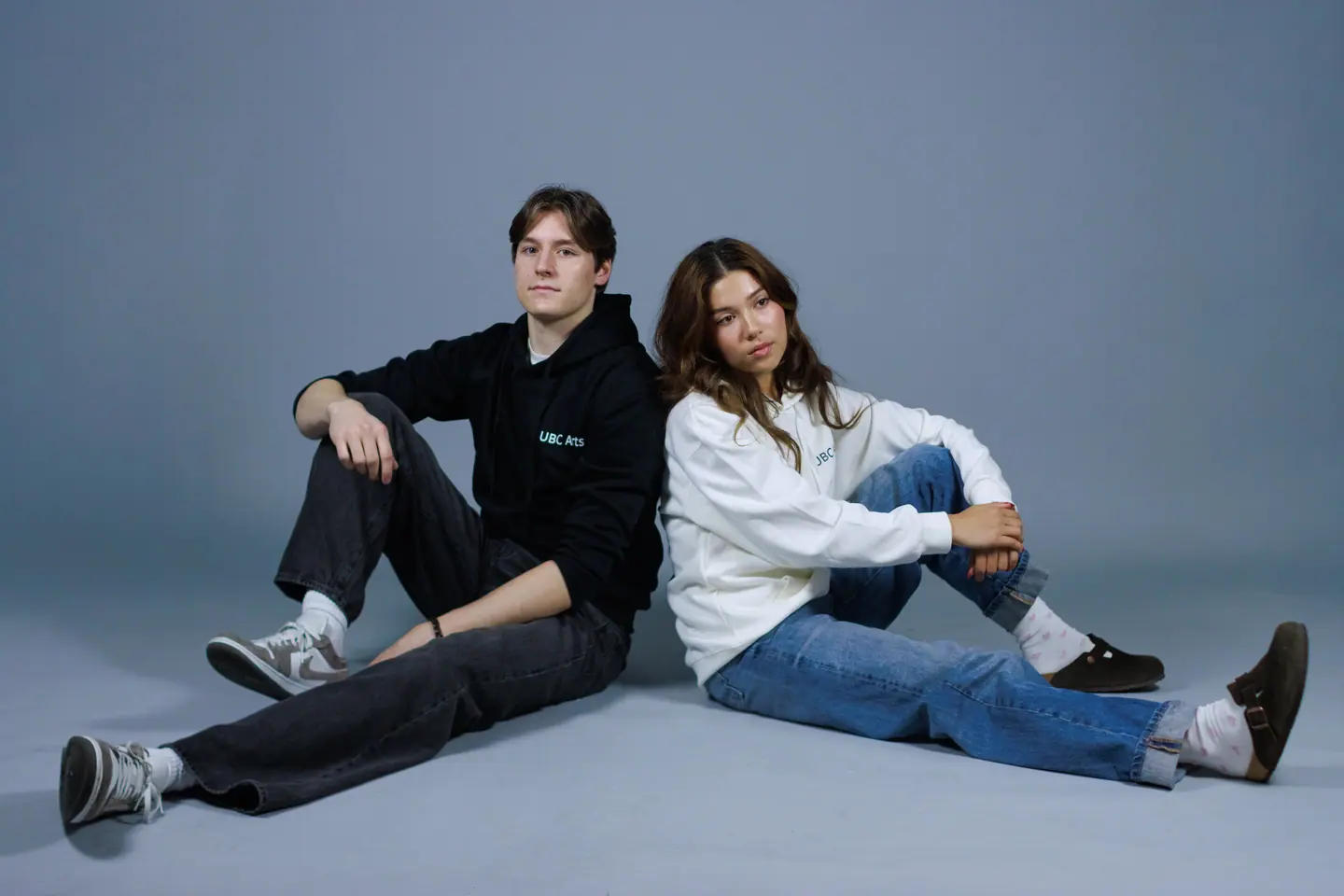 Two students sitting on the floor in UBC Arts tops, one black and one cream